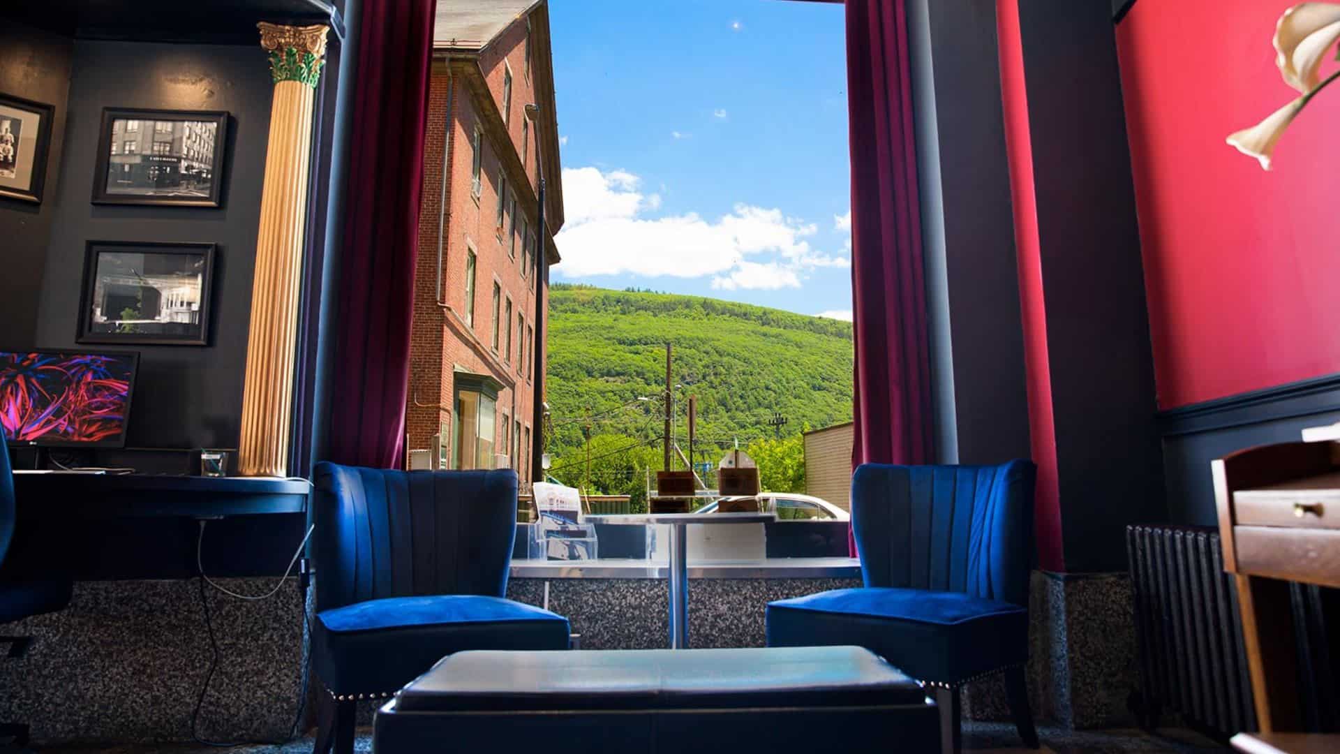 Sitting area with two blue velvet chairs and small table with large window with a view of a green tree covered mountain