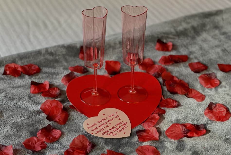 Two red heart shaped Champagne glasses sitting on a red heart tray on top of red flower petals on a gray blanket