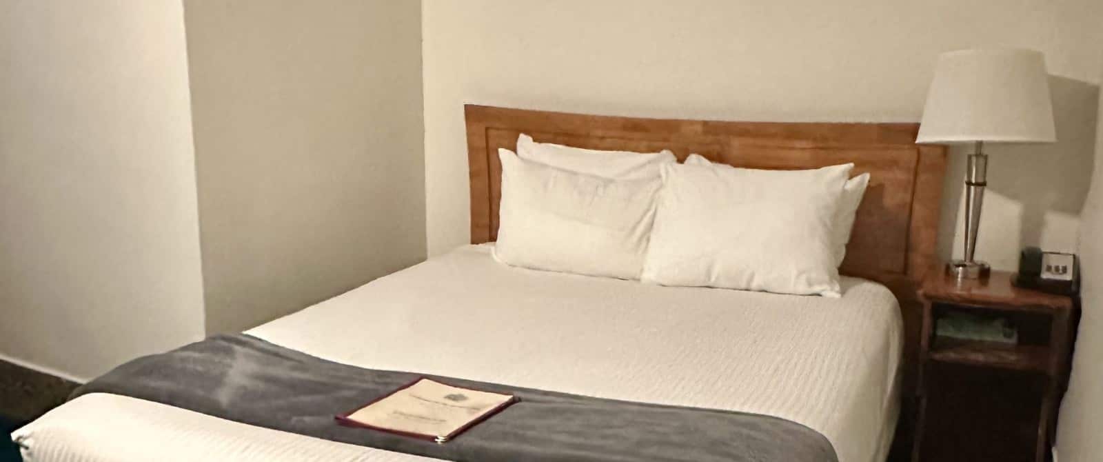 Bedroom with white walls, wooden headboard, white bedding, and nightstand with lamp