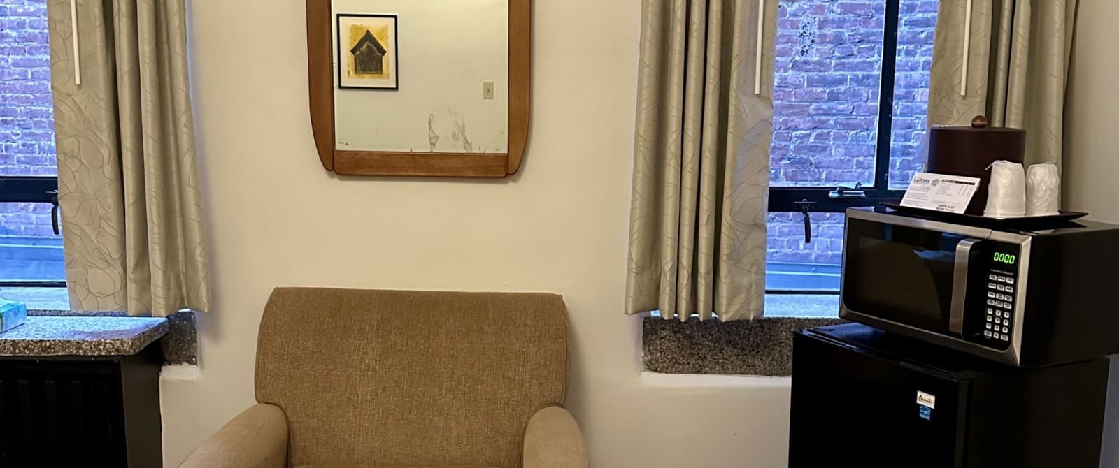 Bedroom with white walls, tan upholstered armchair, microwave, and mini fridge