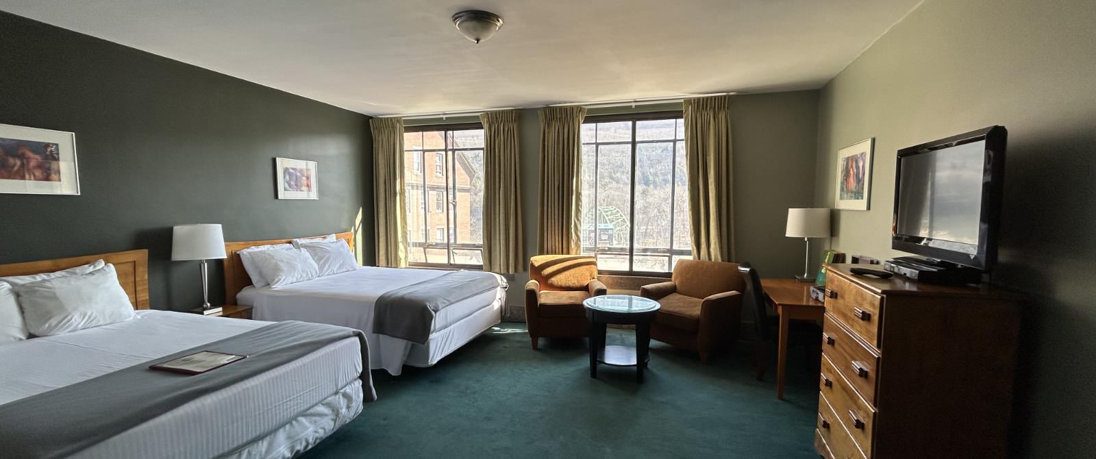 Bedroom with dark gray accent wall, dark green carpeting, two beds each with wooden headboard and white bedding, desk with chair, and sitting area