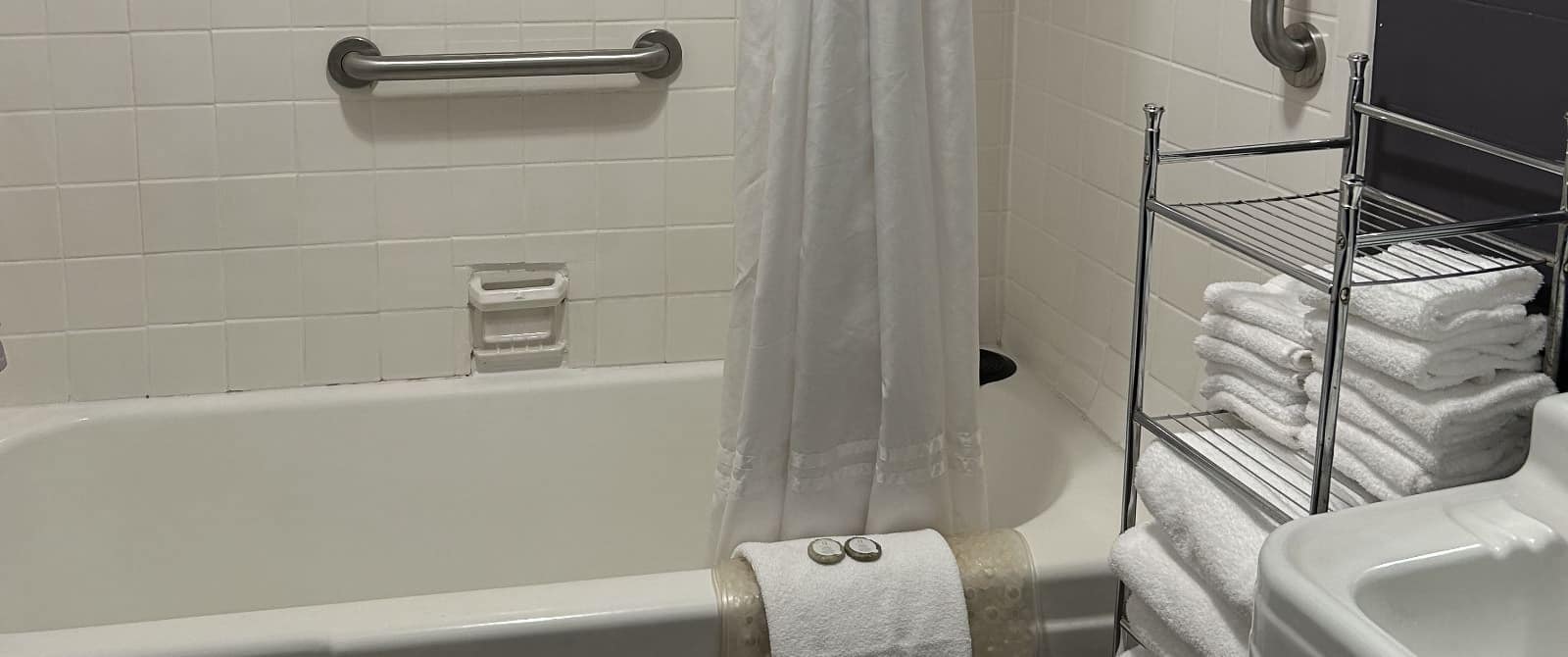 Bathroom with white tiled tub and shower combo, white sink, and silver rack with white towels