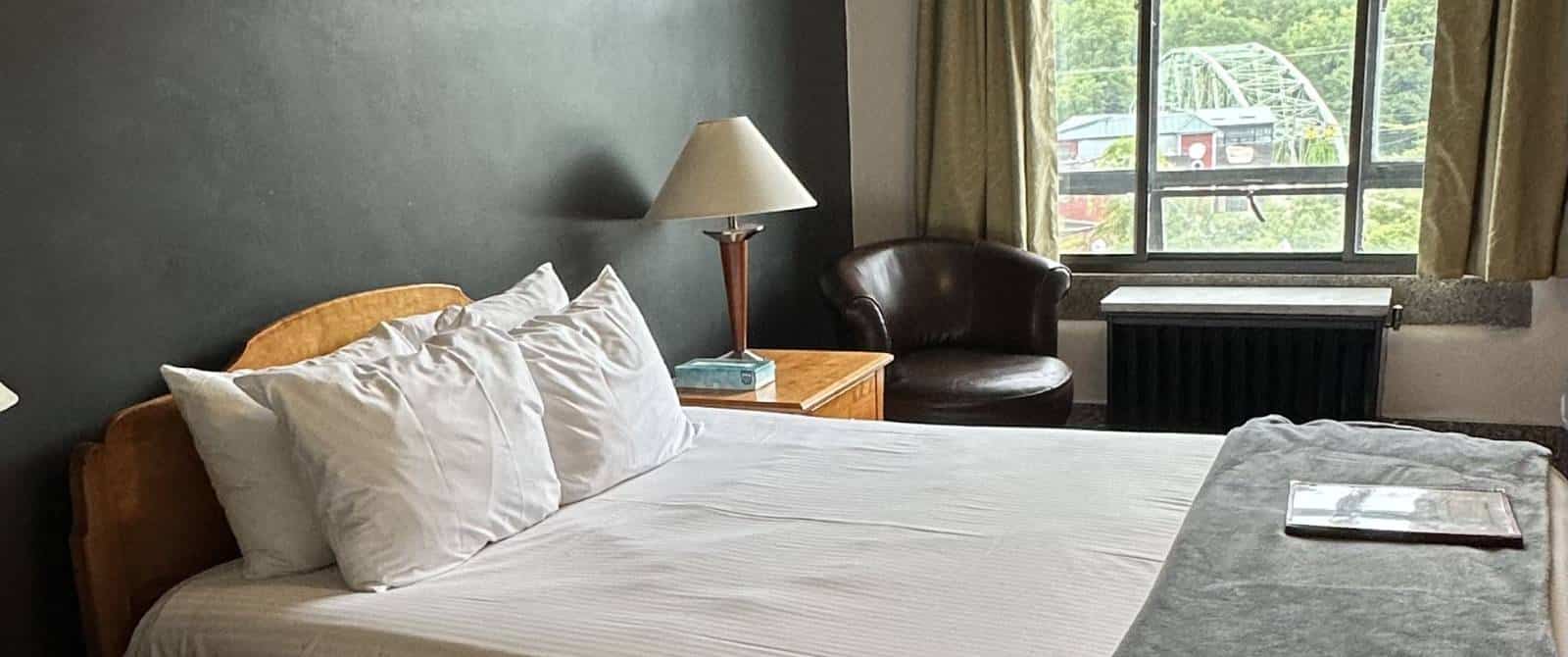 Bedroom with dark gray walls, wooden headboard, white bedding, and wooden nightstand, and black leather chair near window