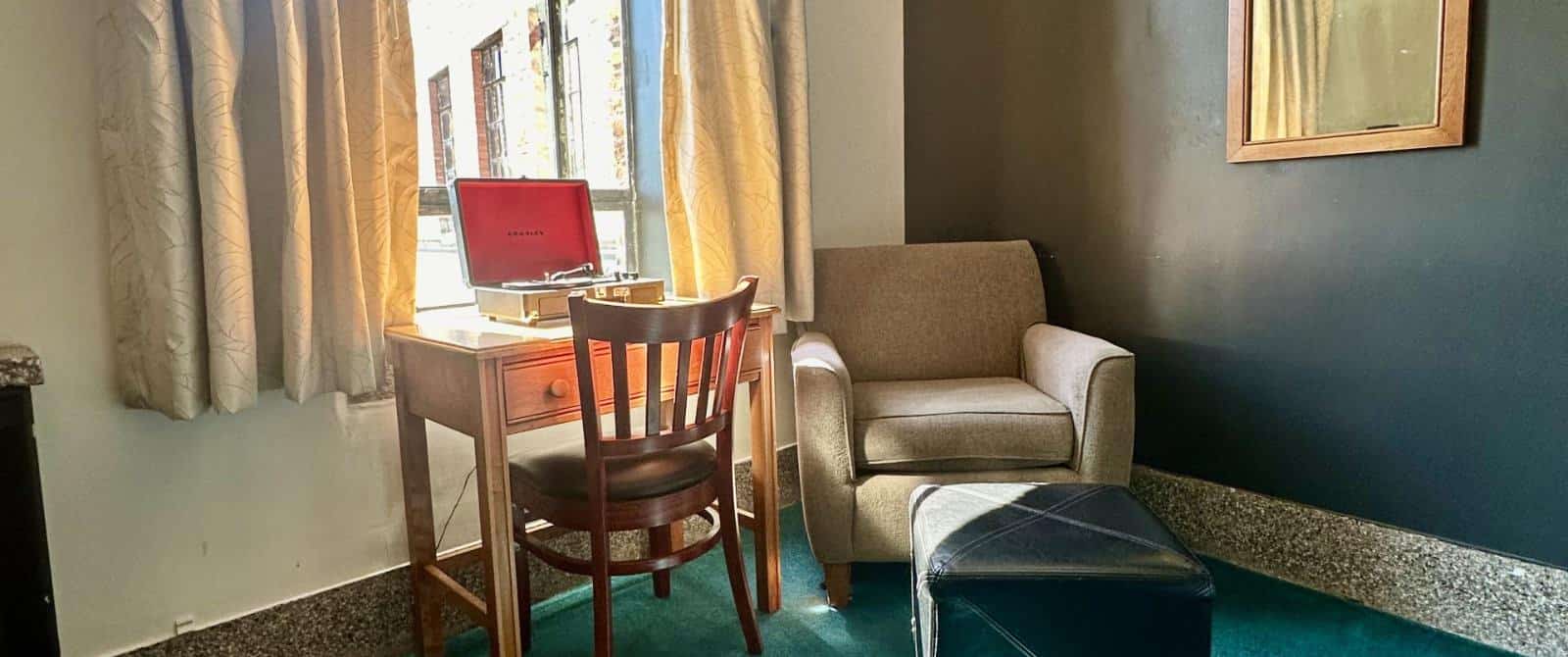 Bedroom with dark gray and white walls, dark emerald carpeting, taupe upholstered armchair, and small desk with chair