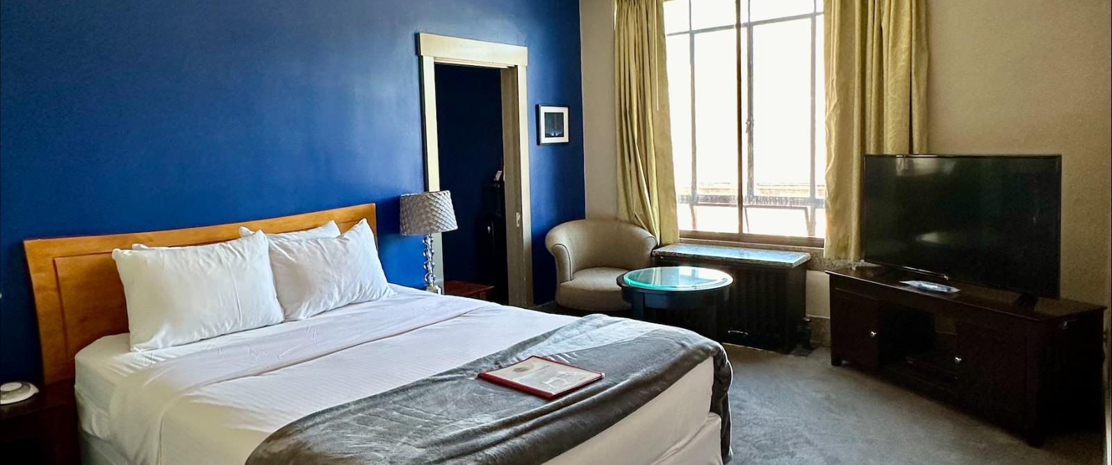 Bedroom with dark blue accent wall, gray carpeting, bed with wooden headboard, white bedding, and sitting area