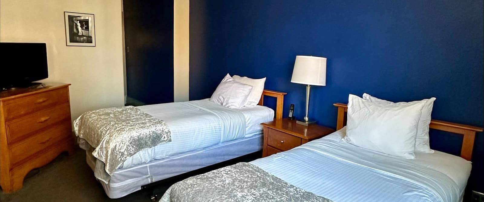 Bedroom with dark blue accent wall, gray carpeting, two beds each with wooden headboard and light blue bedding, nightstand with lamp, and wooden dresser