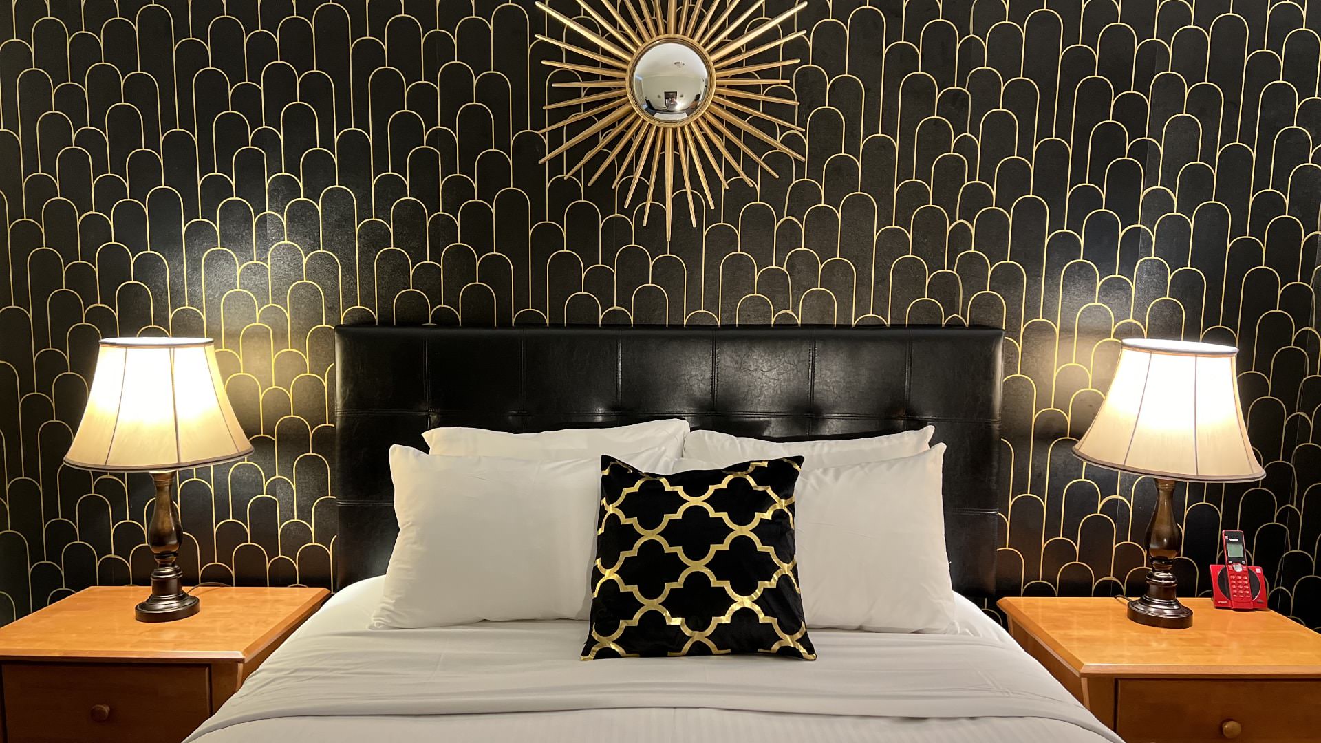 Bedroom with black and gold art deco wallpaper, black leather headboard, white bedding, and wooden nightstands with lamps