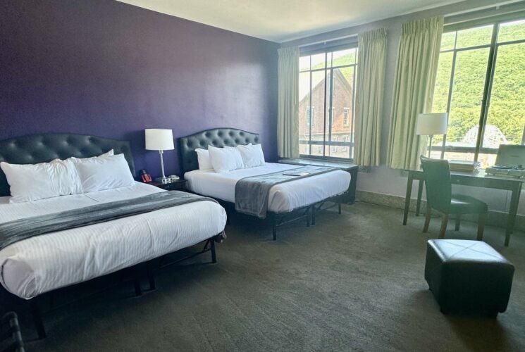 Bedroom with dark purple accent wall, gray carpeting, two beds each with black leather headboard and white bedding, and desk with chair