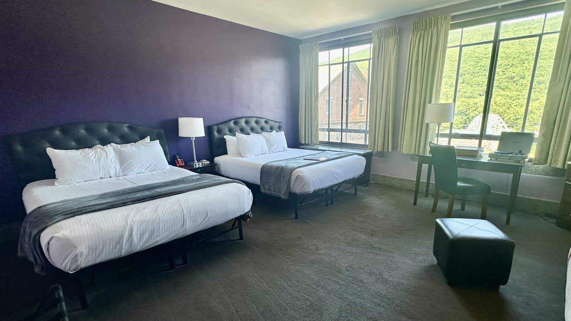 Bedroom with dark purple accent wall, gray carpeting, two beds each with black leather headboard and white bedding, and desk with chair