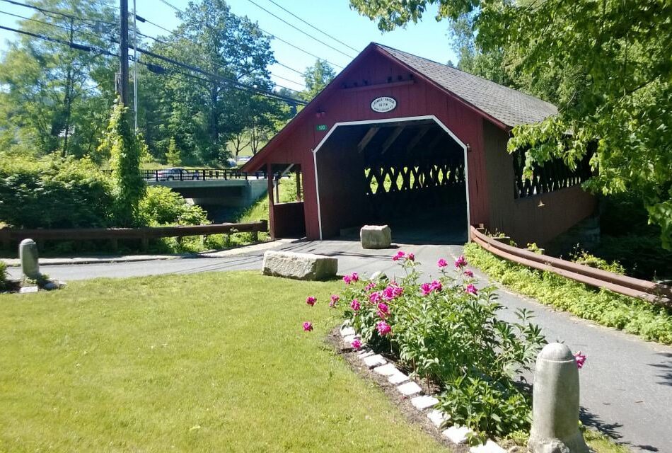 Old Red covered bridge surrounded by green grass and flower bed with pink flowers