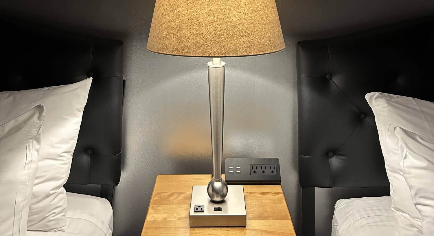 Close up view of wooden nightstand with lamp and charging station between two beds with black leather headboard and white linens